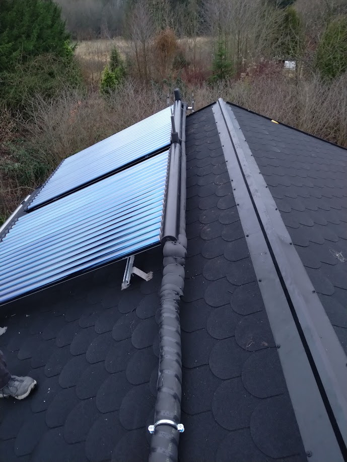 Montaż kolektorów słonecznych na dachu pokrytym gontem bitumicznym, widoczne rury odprowadzające ciepło, fragment buta montera w lewym dolnym rogu.