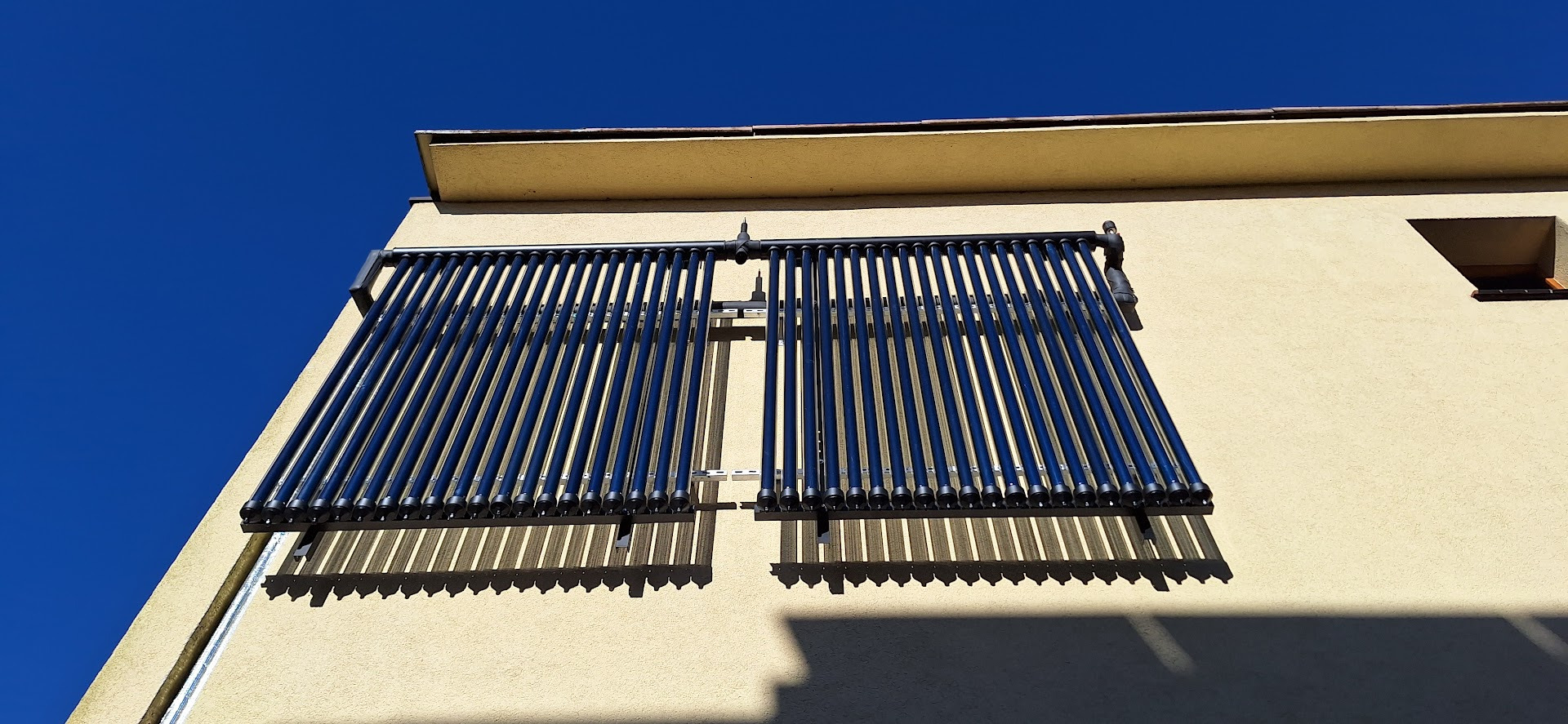 Dwa panele kolektorów słonecznych rurowych zamontowane na fasadzie budynku o jasnej elewacji, widoczne rury i metalowa konstrukcja montażowa, na tle intensywnego błękitu nieba.