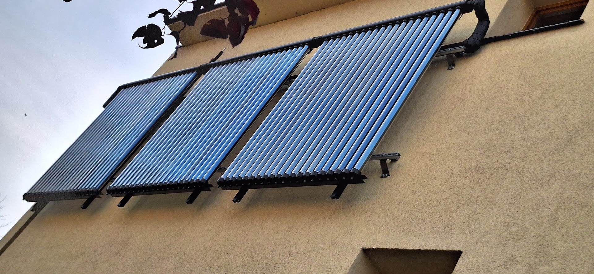 Trzy panele kolektorów słonecznych rurowych zamontowane na elewacji budynku w pochmurny dzień, widoczne czarne rury instalacji i uchwyty mocujące.