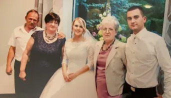 Zdjęcie z uroczystości urodzinowej matki, z widoczną jubilatką w towarzystwie rodziny, w tle dekoracja kwiatowa.