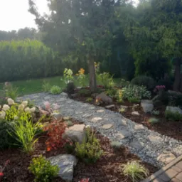 Aranżacja ogrodu z kamienną ścieżką, ozdobnymi kamieniami, korą, różnorodnymi roślinami kwitnącymi i iglakami w tle, oświetlona popołudniowym słońcem.