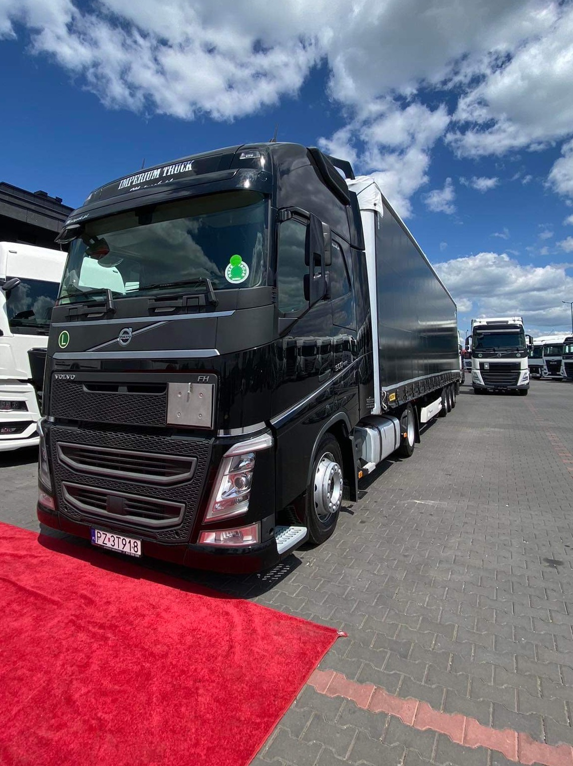 Czarna ciężarówka Volvo FH z naczepą na czerwonym dywanie, tablica rejestracyjna PZ 3T918, błękitne niebo z chmurami w tle.