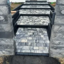 Schody wejściowe do domu wyłożone szaro-białą kostką brukową, obramowane czarnym obrzeżem, z widocznymi słupkami z bloczków betonowych po bokach.