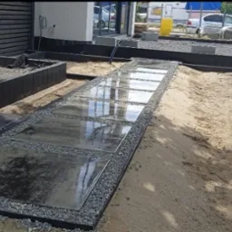 Ścieżka z płyt kamiennych otoczona drobnym żwirem, prowadząca przez piaszczysty teren w Bolesławcu, z widocznymi elementami przygotowania pod dalsze prace.