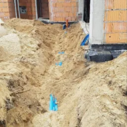 Wykop w piasku pod budynkiem w trakcie budowy, widoczne niebieskie rury instalacyjne i fragment izolacji fundamentów.