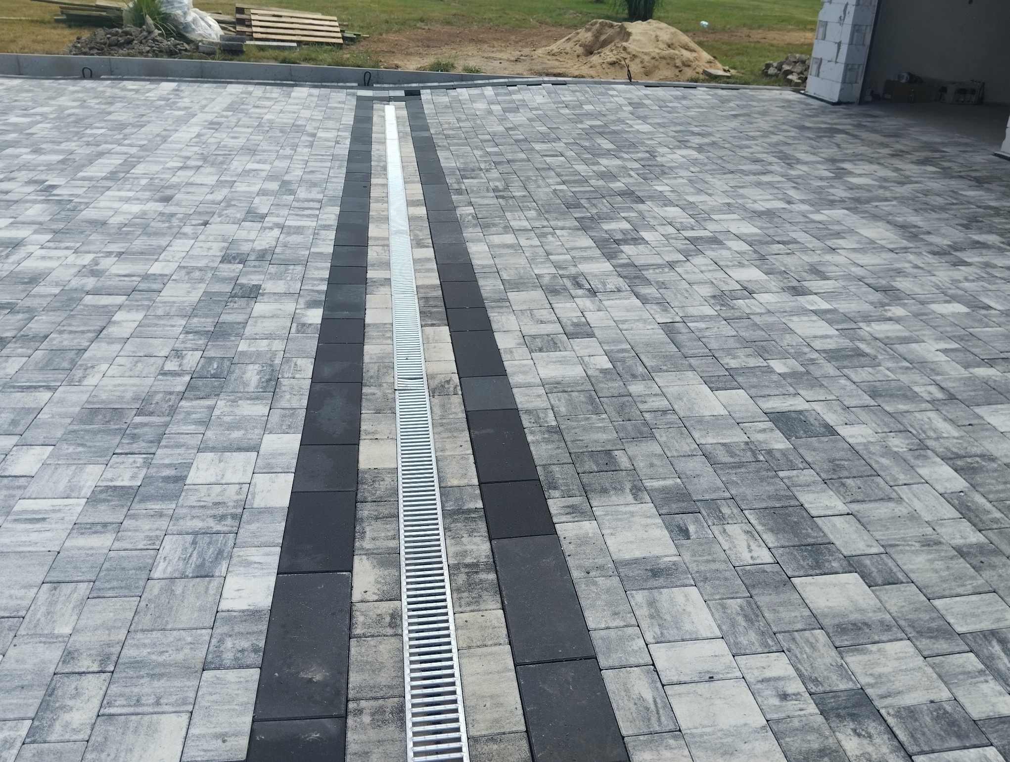 Ułożona nawierzchnia z kostki brukowej w odcieniach szarości, z centralnie umieszczonym odpływem liniowym.