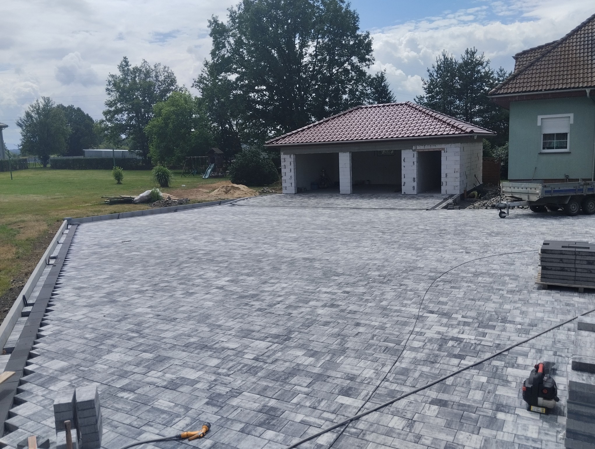 Ułożona szara kostka brukowa na podjeździe z widocznymi krawężnikami i narzędziami, w tle budynek garażu w trakcie budowy i trawnik.