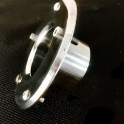 Precyzyjnie wykonany, chromowany element metalowy z czterema śrubami mocującymi, leżący na czarnej, teksturowanej powierzchni.