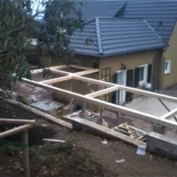 Budowa zadaszenia tarasu z drewnianą konstrukcją szkieletową, widok z góry na plac budowy, w tle dom jednorodzinny z ciemną dachówką.