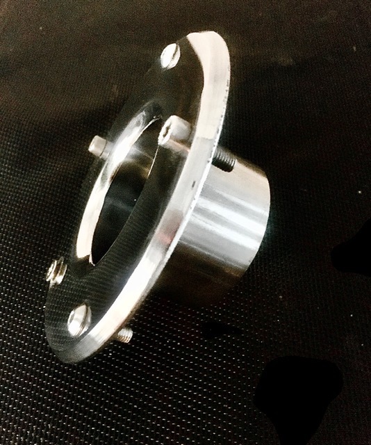 Precyzyjnie wykonany, chromowany element metalowy z czterema śrubami mocującymi, leżący na czarnej, teksturowanej powierzchni.