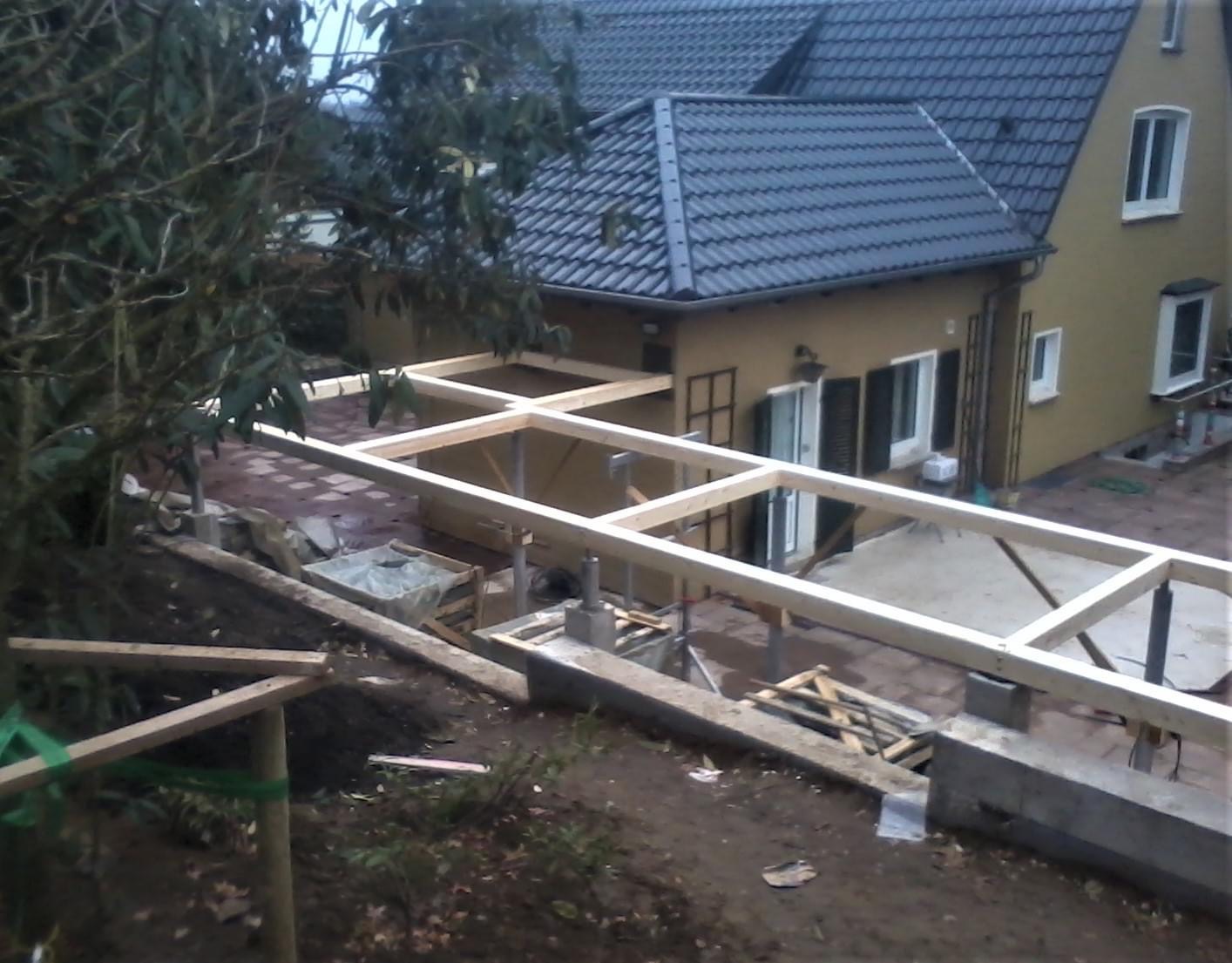 Budowa zadaszenia tarasu z drewnianą konstrukcją szkieletową, widok z góry na plac budowy, w tle dom jednorodzinny z ciemną dachówką.