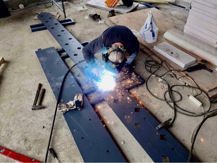 Pracownik w ochronie słuchu i masce spawalniczej łączy elementy metalowe metodą spawania elektrycznego; iskry i dym unoszą się nad spawanym materiałem, widoczne narzędzia i elementy konstrukcji...