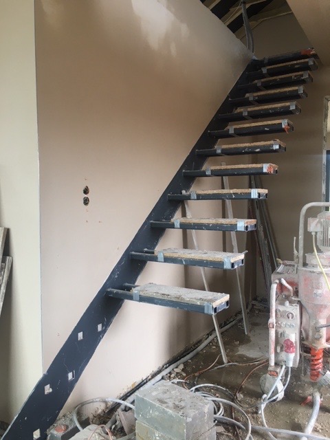 Surowe schody metalowe z czarną konstrukcją i drewnianymi stopniami w trakcie montażu, widoczny sprzęt budowlany i niedokończone ściany.