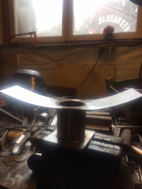 Metalowa konstrukcja z okrągłym otworem, spawana do podstawy, widoczna w warsztacie ślusarskim z narzędziami i elementami metalowymi w tle.