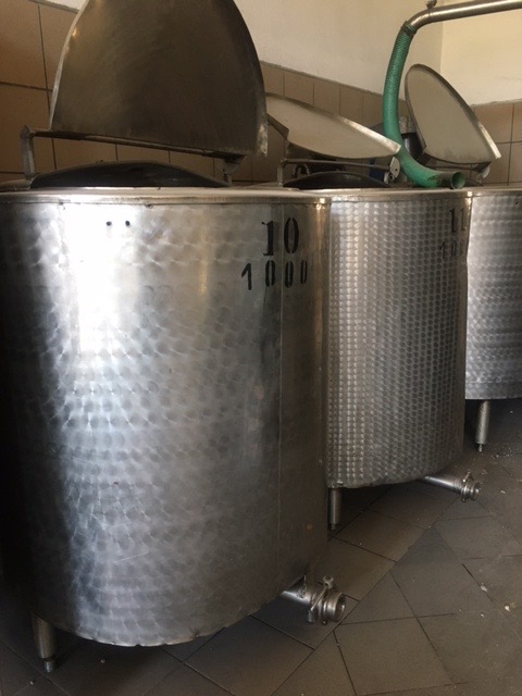 Rzędy dużych, metalowych zbiorników z otwartymi pokrywami, oznaczonych numerami '10' i '1000', stojących na małych nóżkach w pomieszczeniu z kafelkami.