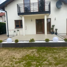 Nowoczesny dom z białymi ścianami, balkonem z czarną balustradą i starannie zagospodarowanym terenem przed wejściem, gdzie widać podświetlane kule dekoracyjne, białe kamienie i małe krzewy.