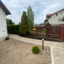 Nowo wybrukowana ścieżka z kostki brukowej w ogrodzie z lampami solarnymi, świeżo posadzone tuje i trawnik w trakcie zakładania, widoczny drewniany płot i brama wjazdowa.