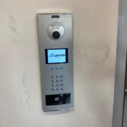 Panel domofonowy FERMAX z wyświetlaczem LCD i klawiaturą numeryczną, zamontowany na surowej, szpachlowanej ścianie. Na ekranie widoczne logo firmy L Elektro Grzegorz Leszczyński.