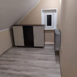 Pokój na poddaszu z panelami podłogowymi imitującymi jasne drewno, beżowymi ścianami, grzejnikiem i szafką z ciemnobrązowym blatem pod oknem.