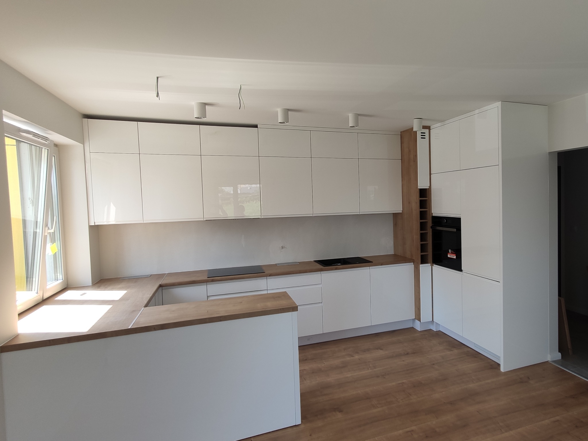 Jasna kuchnia z białymi, lakierowanymi szafkami i drewnianym blatem, widoczne płyty indukcyjne, oświetlenie sufitowe punktowe i podłoga imitująca drewno.