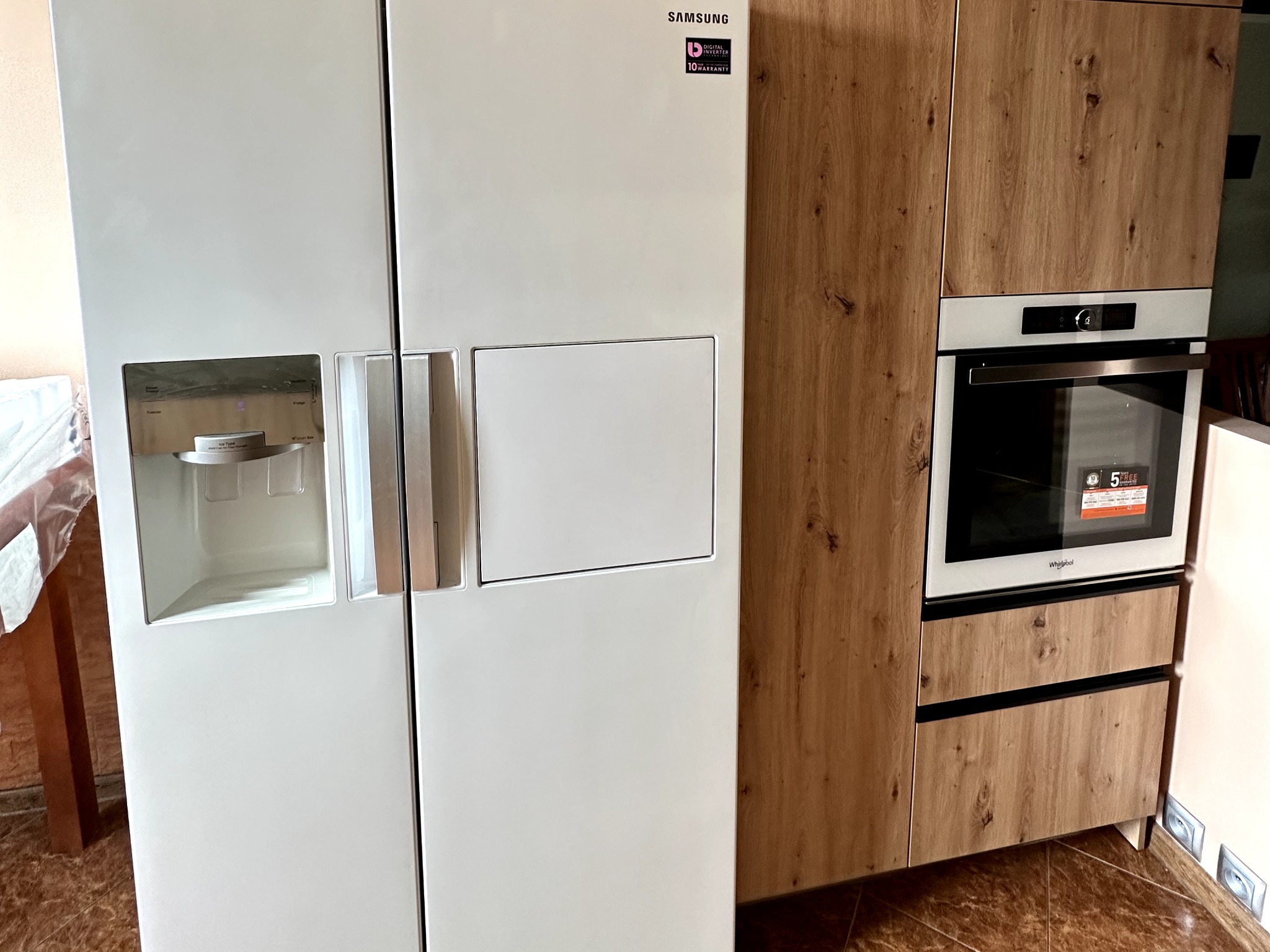 Biała lodówka side-by-side Samsung z dystrybutorem wody i kostkarką, sąsiadująca z zabudową kuchenną z drewnopodobnymi frontami, w której umieszczono piekarnik Whirlpool.