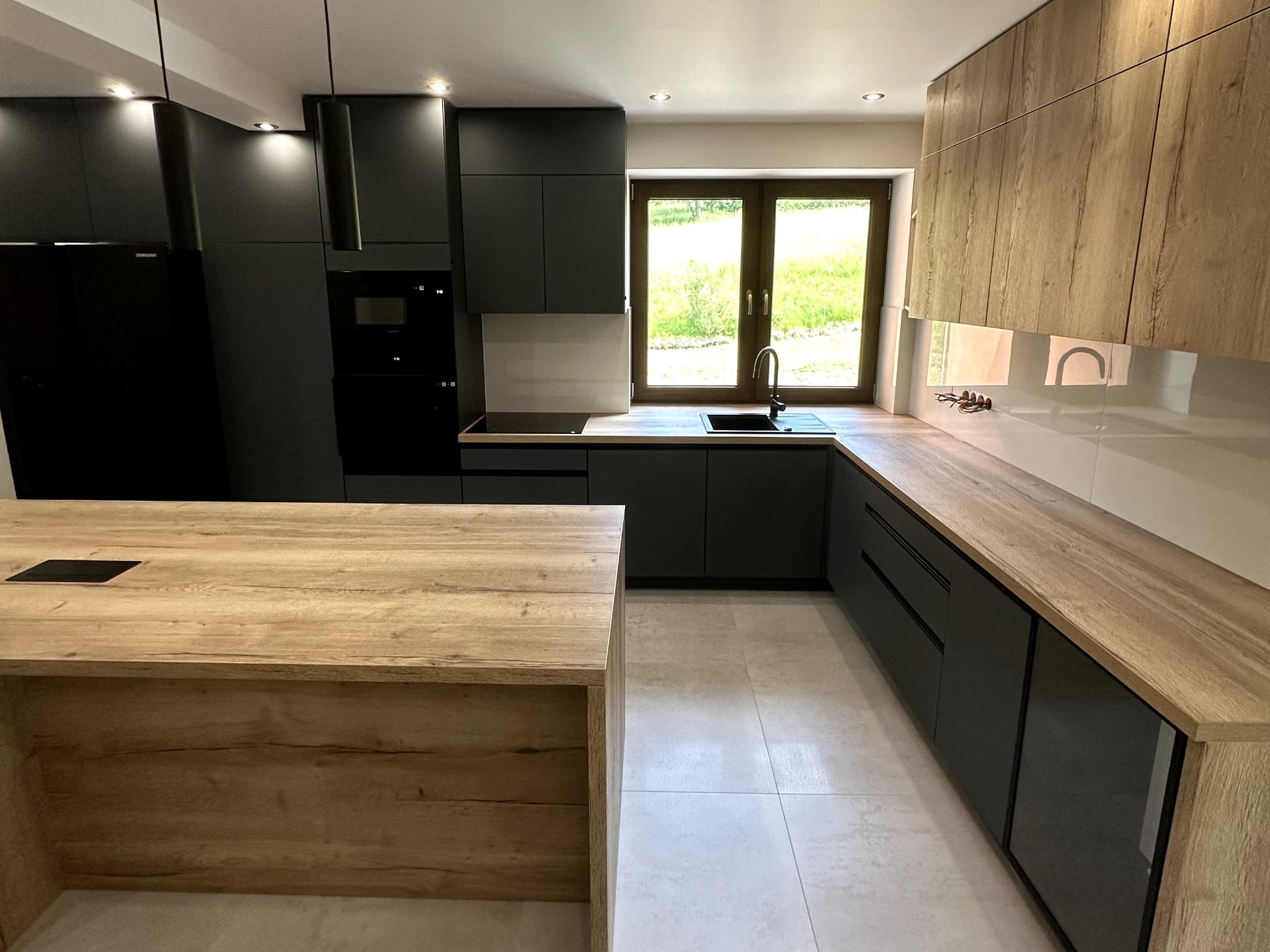 Nowoczesna kuchnia z zabudową łączącą matową czerń i drewnopodobne blaty, wyspa kuchenna z gniazdem elektrycznym w blacie, widok na okno z zielenią za szybą.