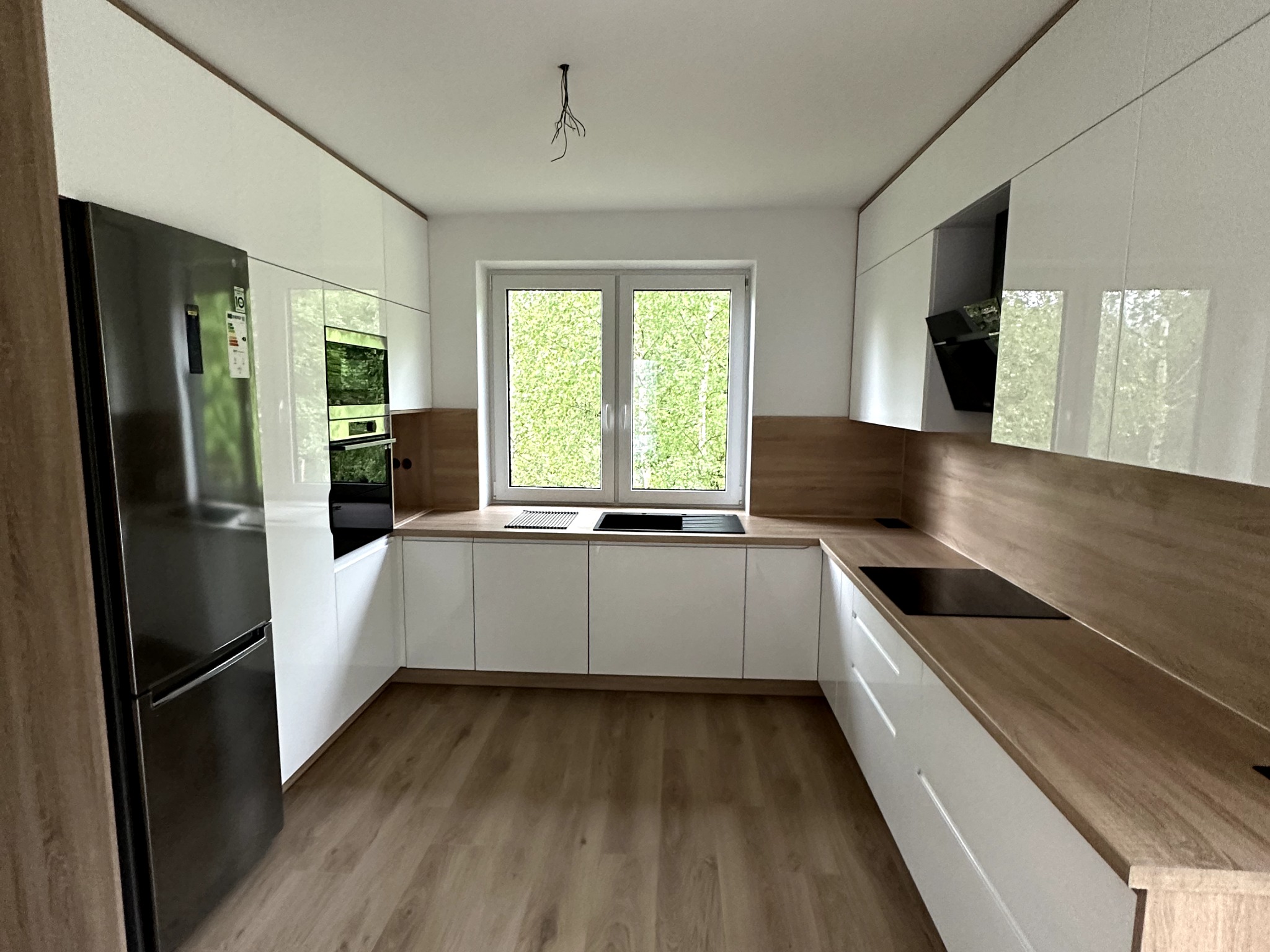 Nowoczesna kuchnia z białymi, lakierowanymi frontami szafek i drewnopodobnym blatem, z widocznym oknem i wystającymi kablami oświetleniowymi.