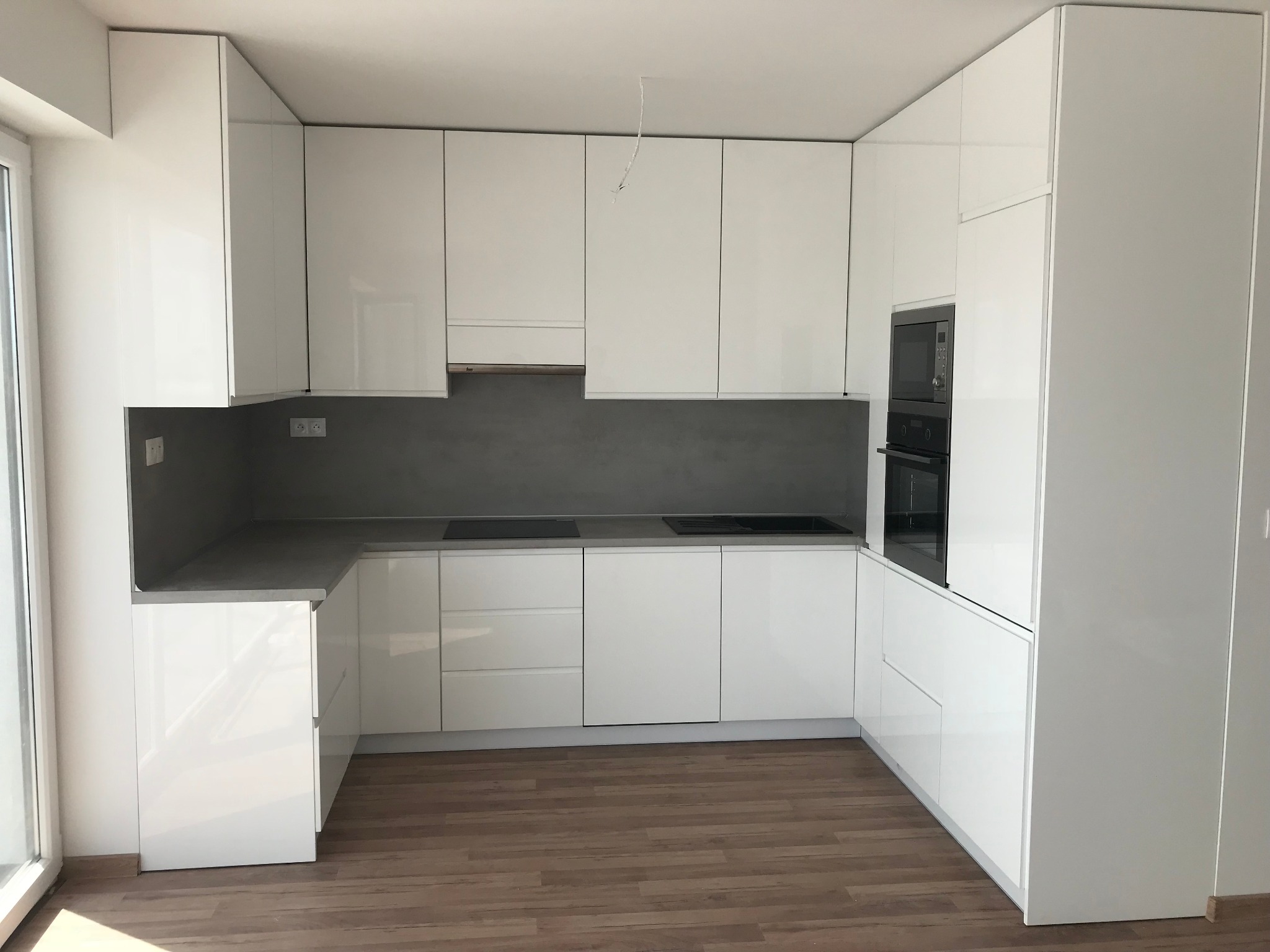 Nowoczesna, minimalistyczna kuchnia z białymi, lakierowanymi frontami, szarym blatem i drewnopodobną podłogą, z wbudowanym piekarnikiem i płytą grzewczą.