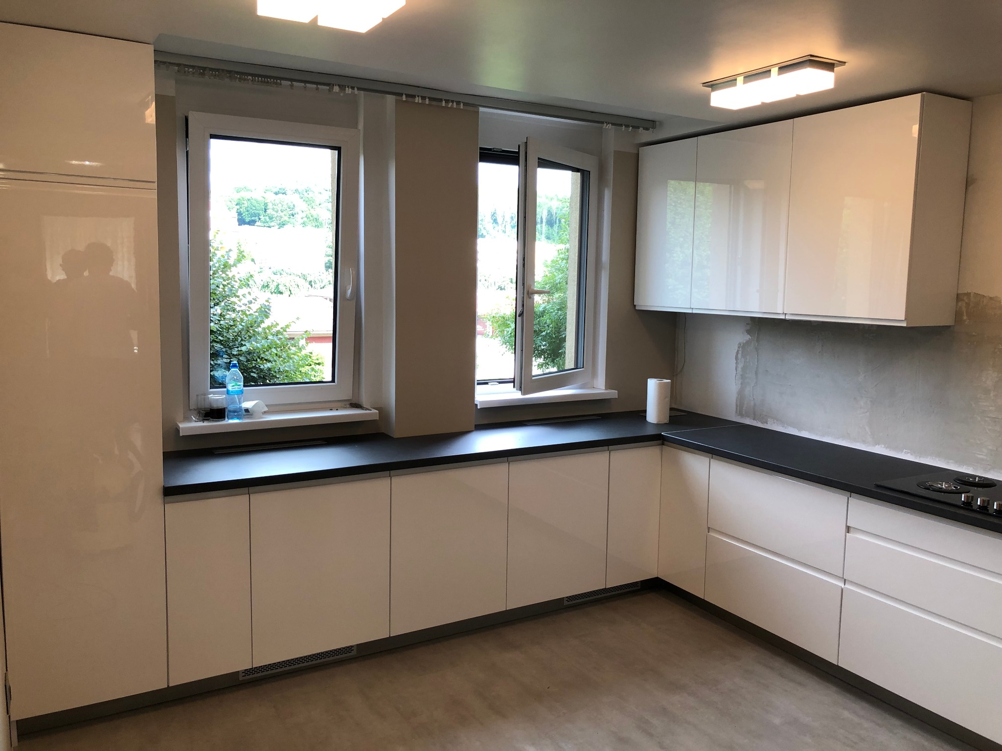 Nowoczesna kuchnia z białymi, lakierowanymi frontami szafek i czarnym blatem, widok na okno z zielenią na zewnątrz, ściana częściowo w stanie surowym.