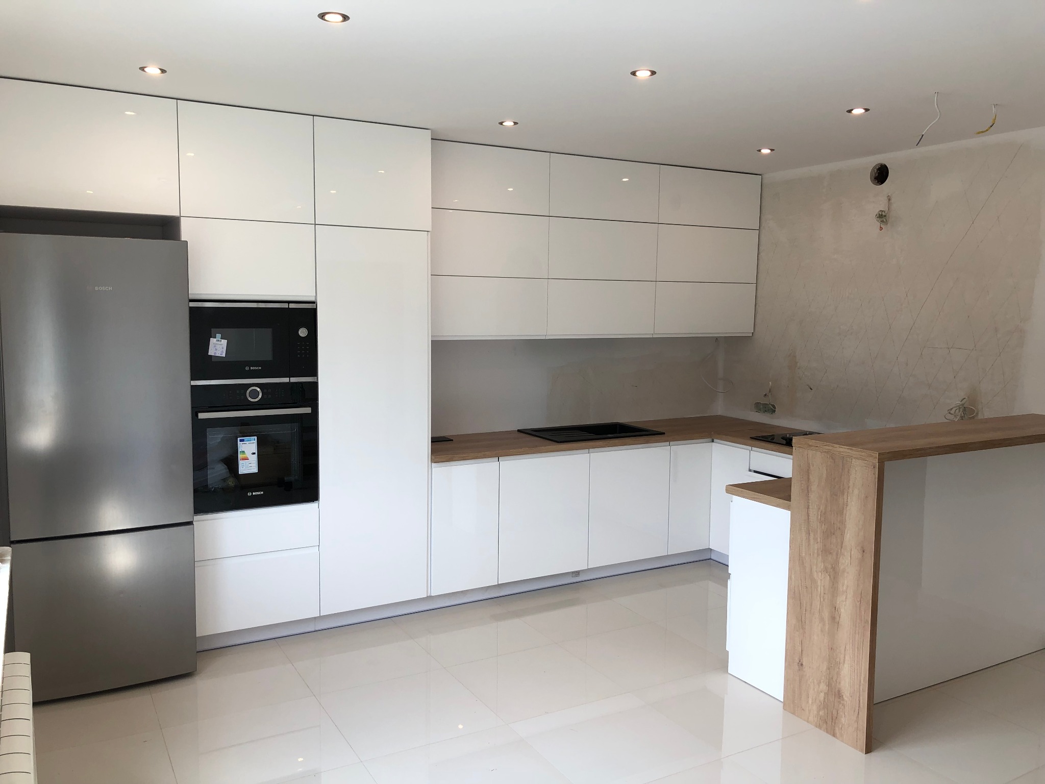 Nowoczesna, biała kuchnia z połyskującymi frontami, drewnianym blatem i barkiem, oraz srebrną lodówką Bosch, widziana z perspektywy podłogi, z elementami wykończeniowymi ścian w tle.
