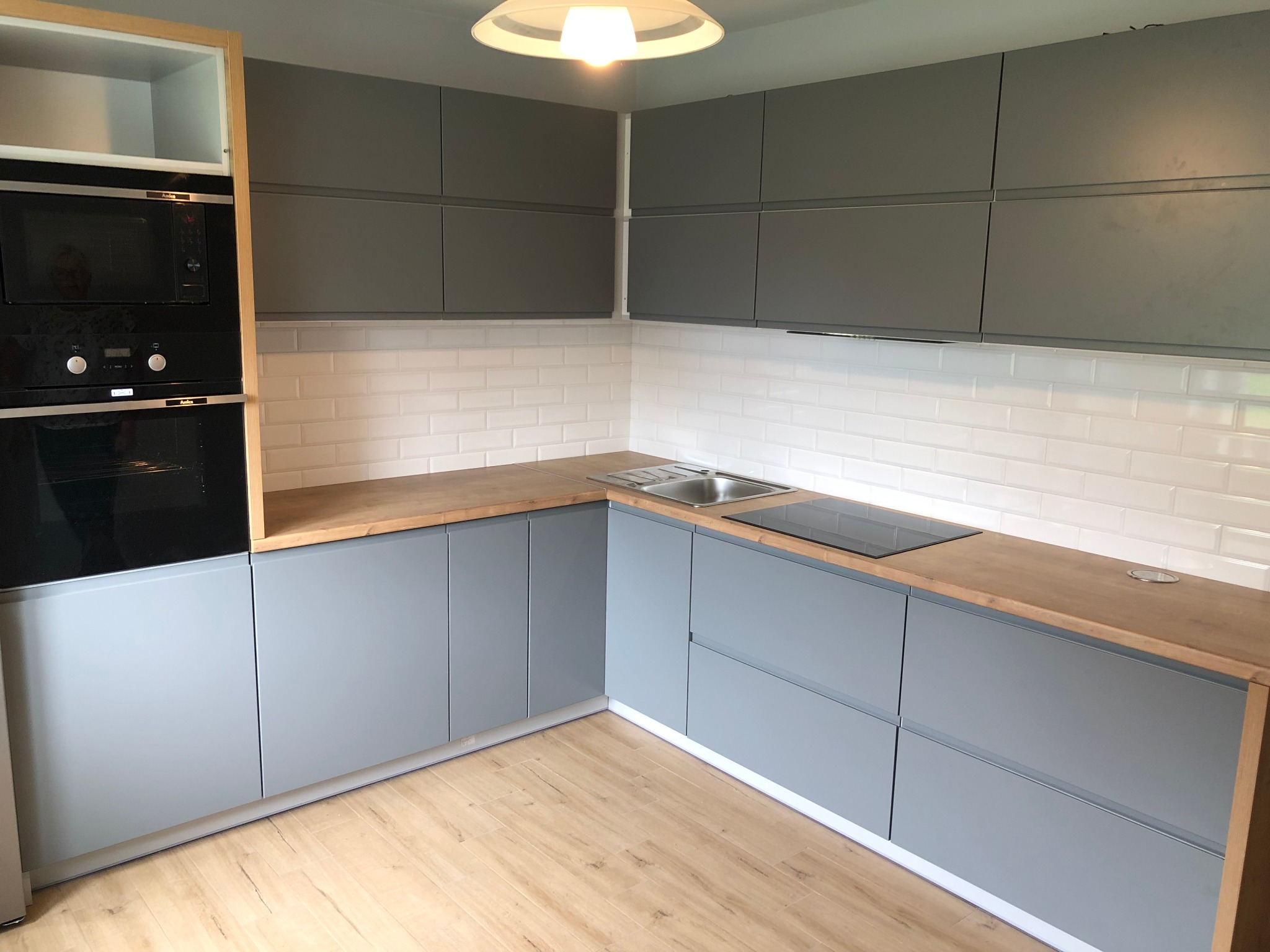 Szare szafki kuchenne z drewnianym blatem, białe kafelki na ścianie, płyta indukcyjna i zlew ze stali nierdzewnej, oświetlenie sufitowe.