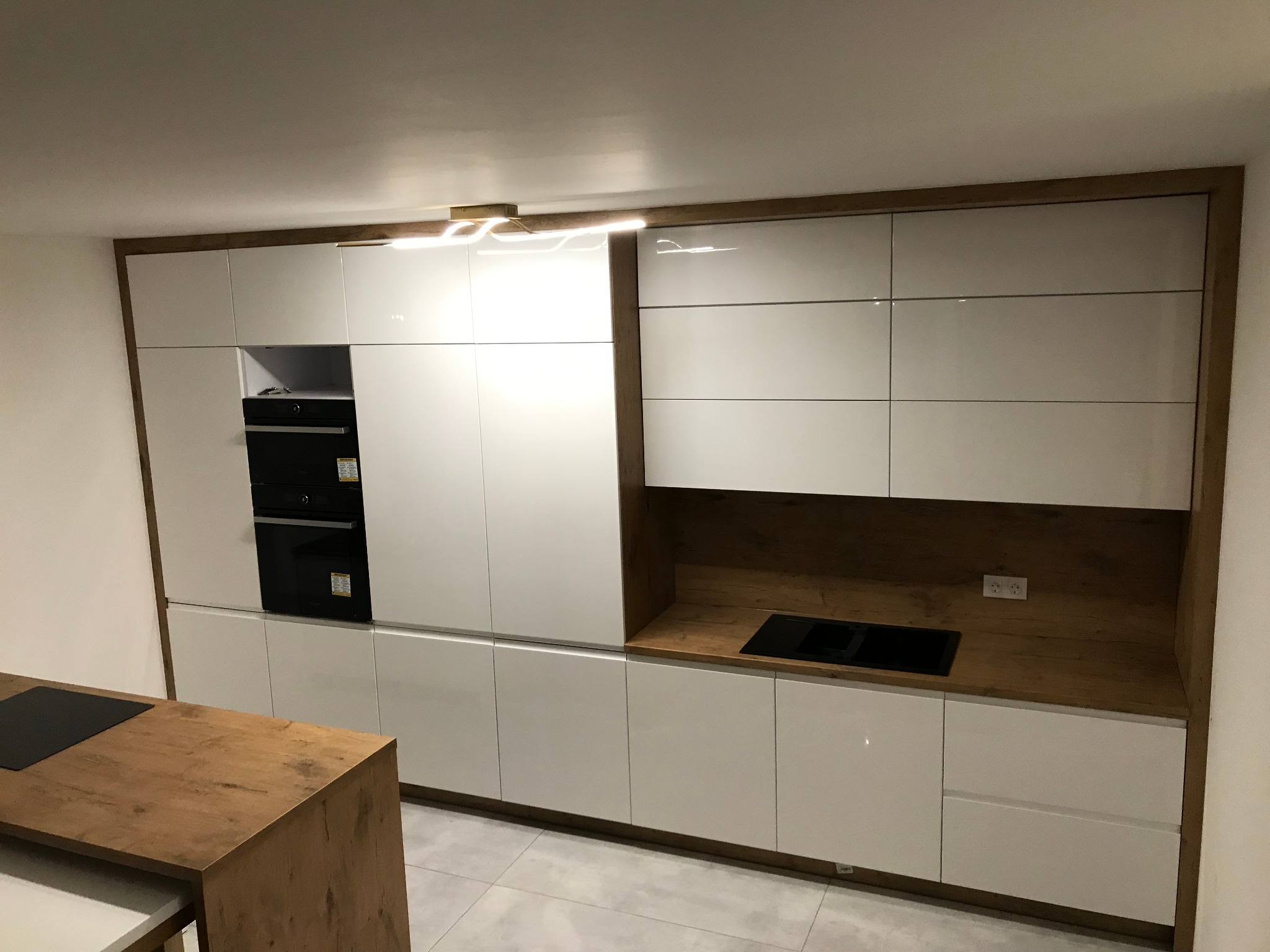 Nowoczesna kuchnia z białymi, lakierowanymi frontami szafek i drewnianymi elementami wykończeniowymi, wbudowany piekarnik i płyta indukcyjna, oświetlenie podszafkowe.