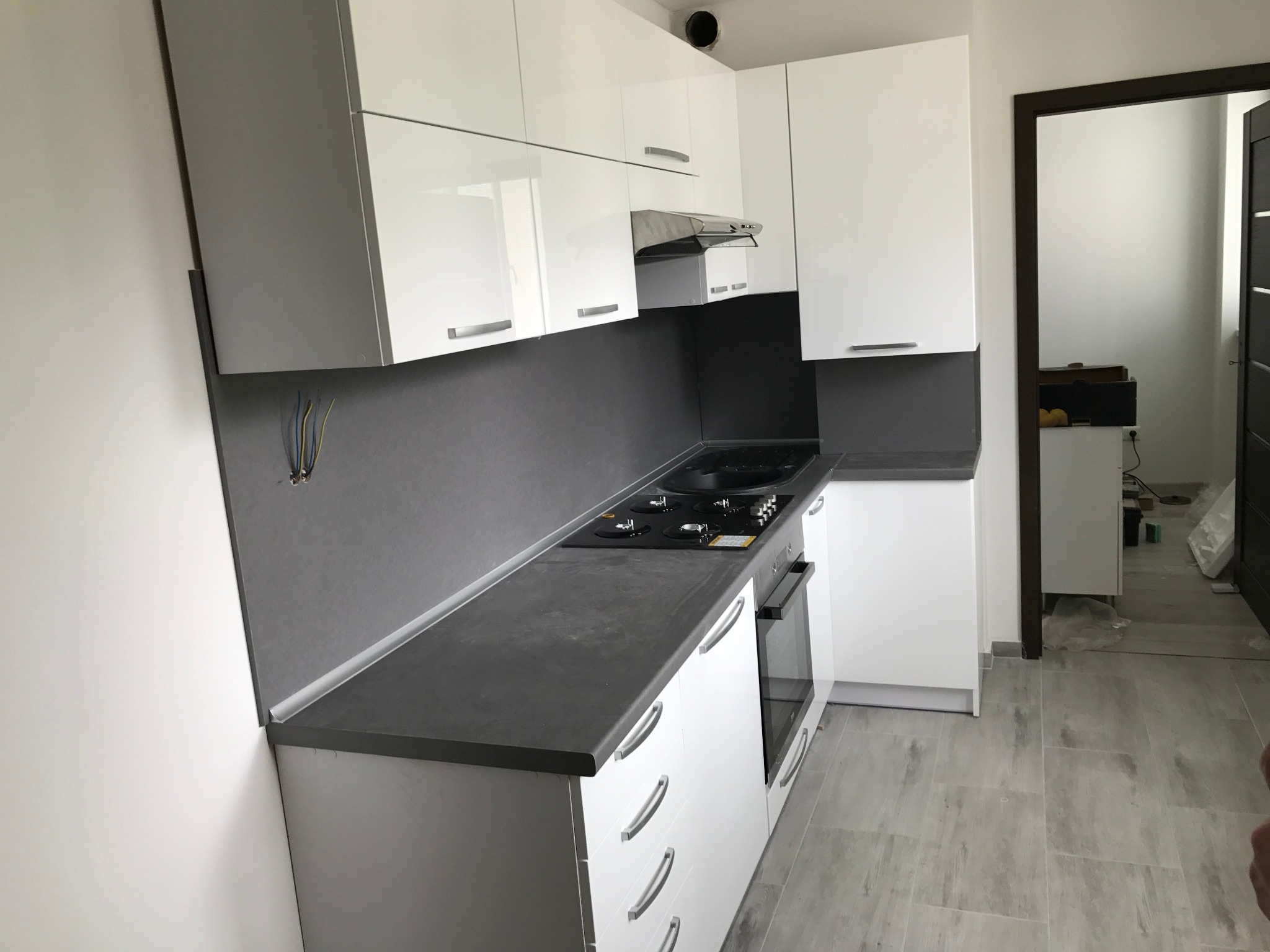 Nowoczesna kuchnia z białymi lakierowanymi frontami, szarym blatem i płytkami, widoczne niedokończone podłączenie elektryczne.