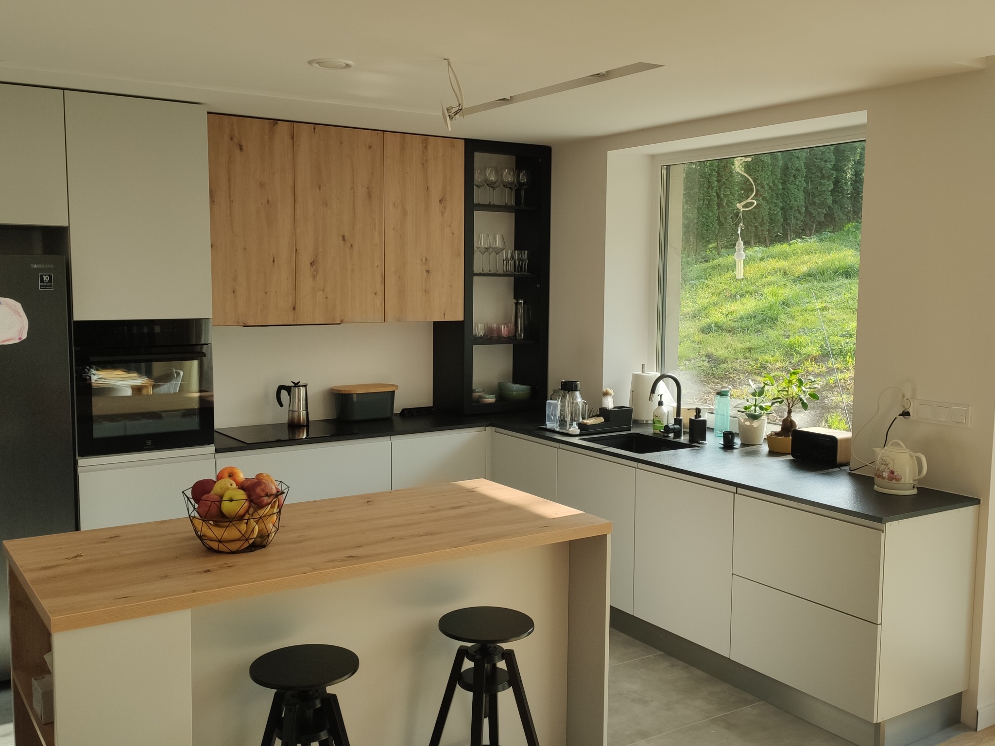 Jasna kuchnia z drewnianym blatem wyspy, czarnymi blatami roboczymi i widokiem na zieleń za oknem.
