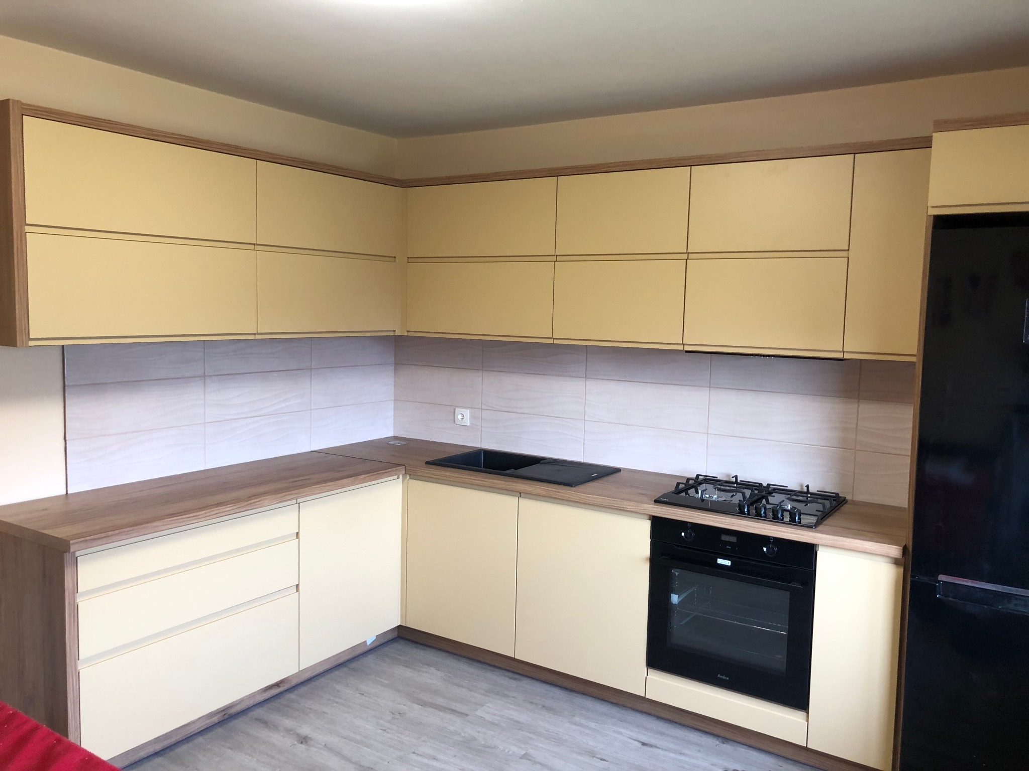 Jasna kuchnia z beżowymi frontami szafek i drewnianymi blatami, czarna płyta gazowa i piekarnik w zabudowie, lodówka w kolorze czarnym po prawej stronie.