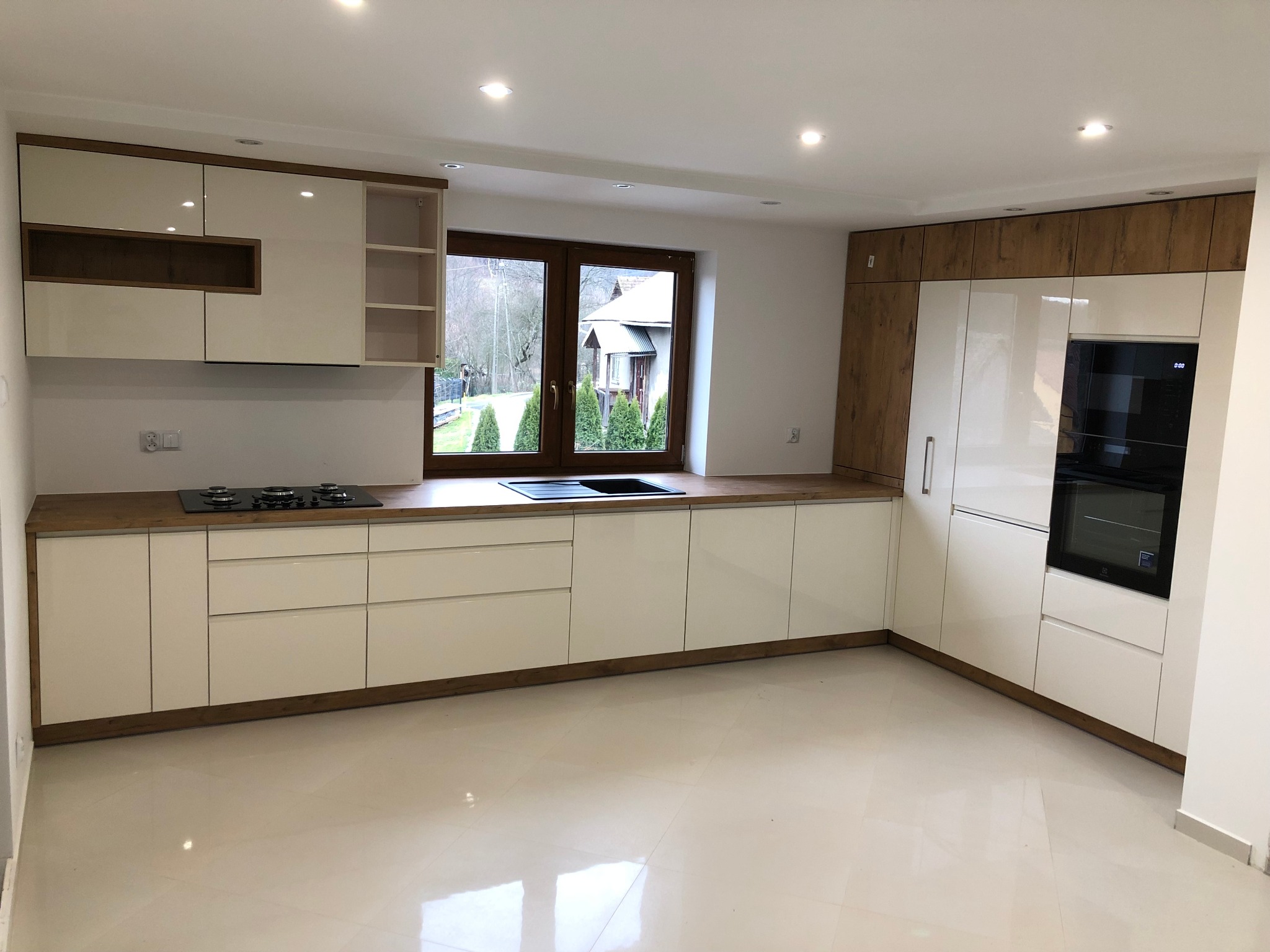 Nowoczesna kuchnia z białymi lakierowanymi frontami, drewnianym blatem i zabudową do sufitu, widok na okno z zielonym ogrodem.