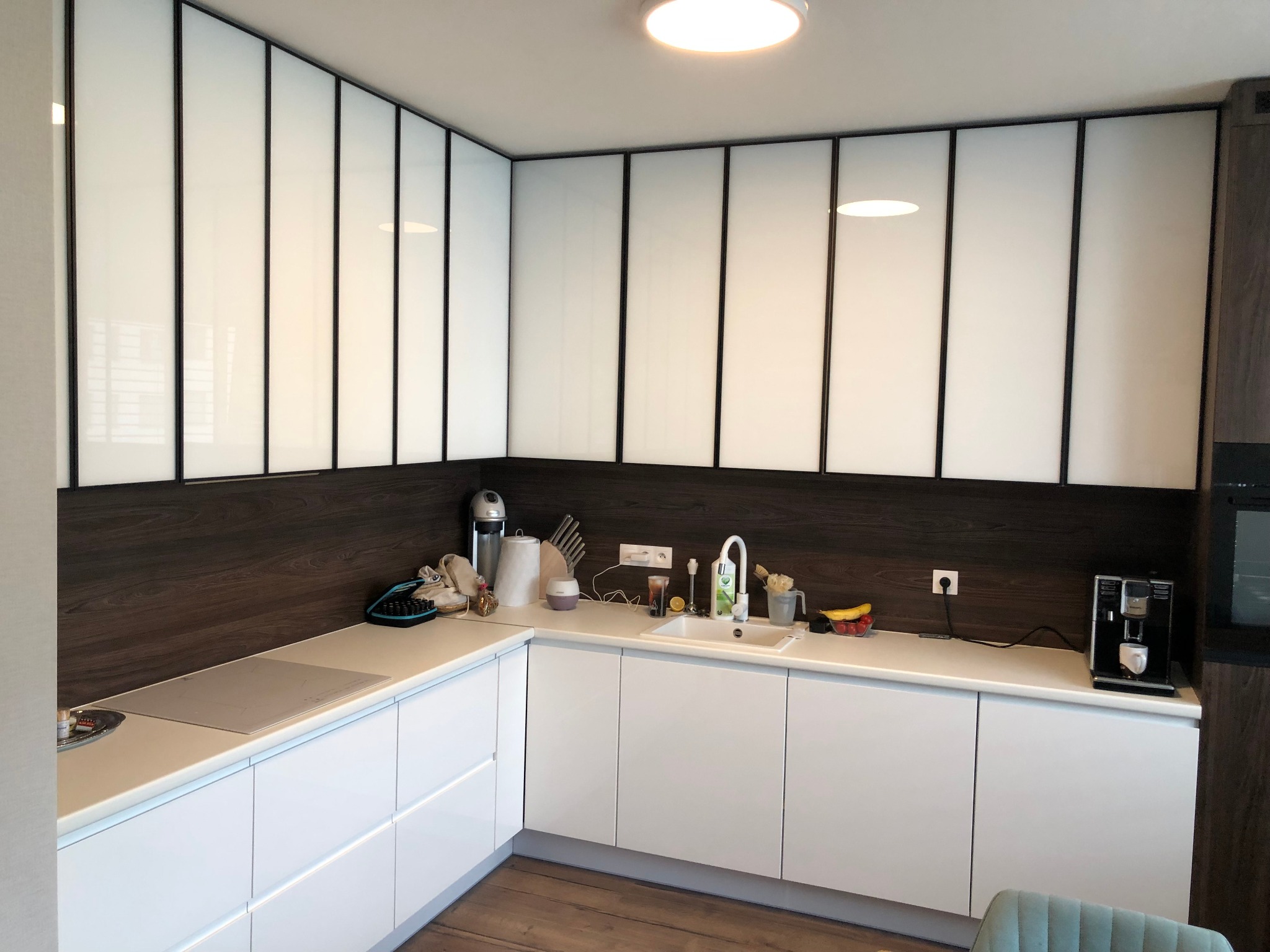 Nowoczesna kuchnia z białymi, gładkimi szafkami dolnymi i górnymi wykończonymi panelami z czarnymi obramowaniami, drewnopodobny panel nad blatem, ekspres do kawy i owoce na blacie.