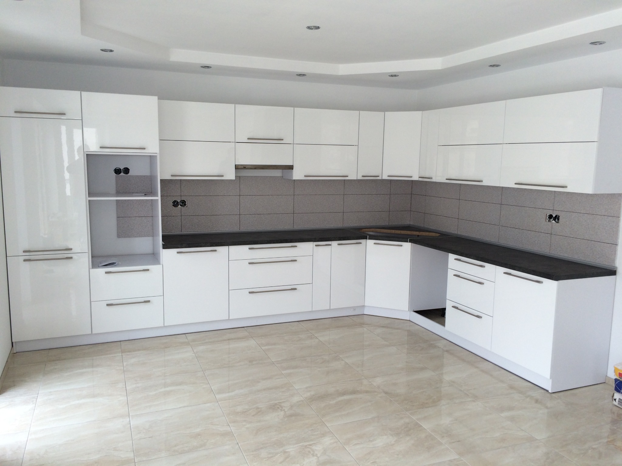 Nowoczesna, biała kuchnia z błyszczącymi frontami, ciemnym blatem i szarymi płytkami nad blatem, widoczne gniazdka elektryczne w ścianie, oświetlenie sufitowe punktowe.