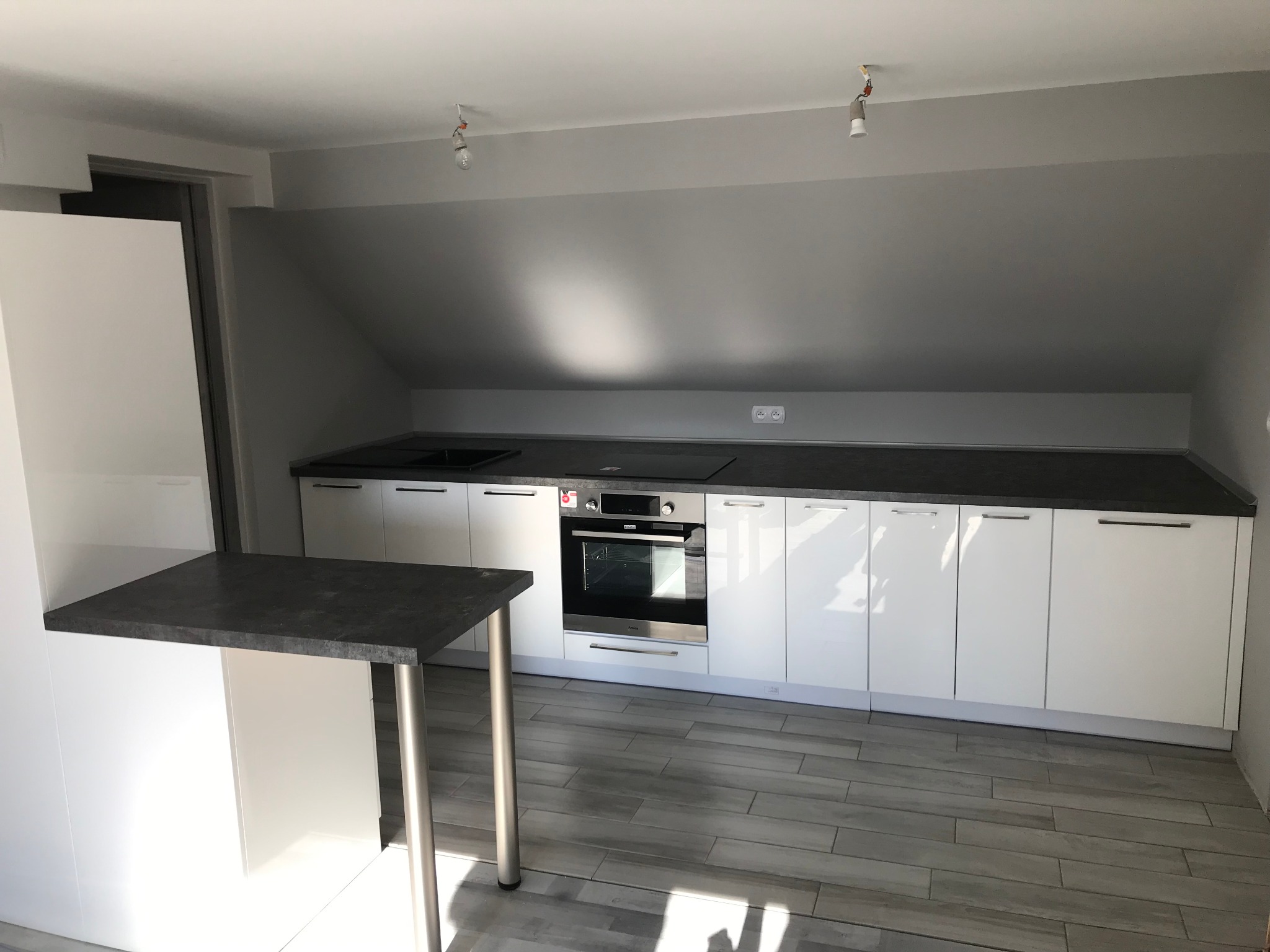 Nowoczesna biała kuchnia w skosie z ciemnym blatem, wyspą i płytą indukcyjną, oświetlenie tymczasowe