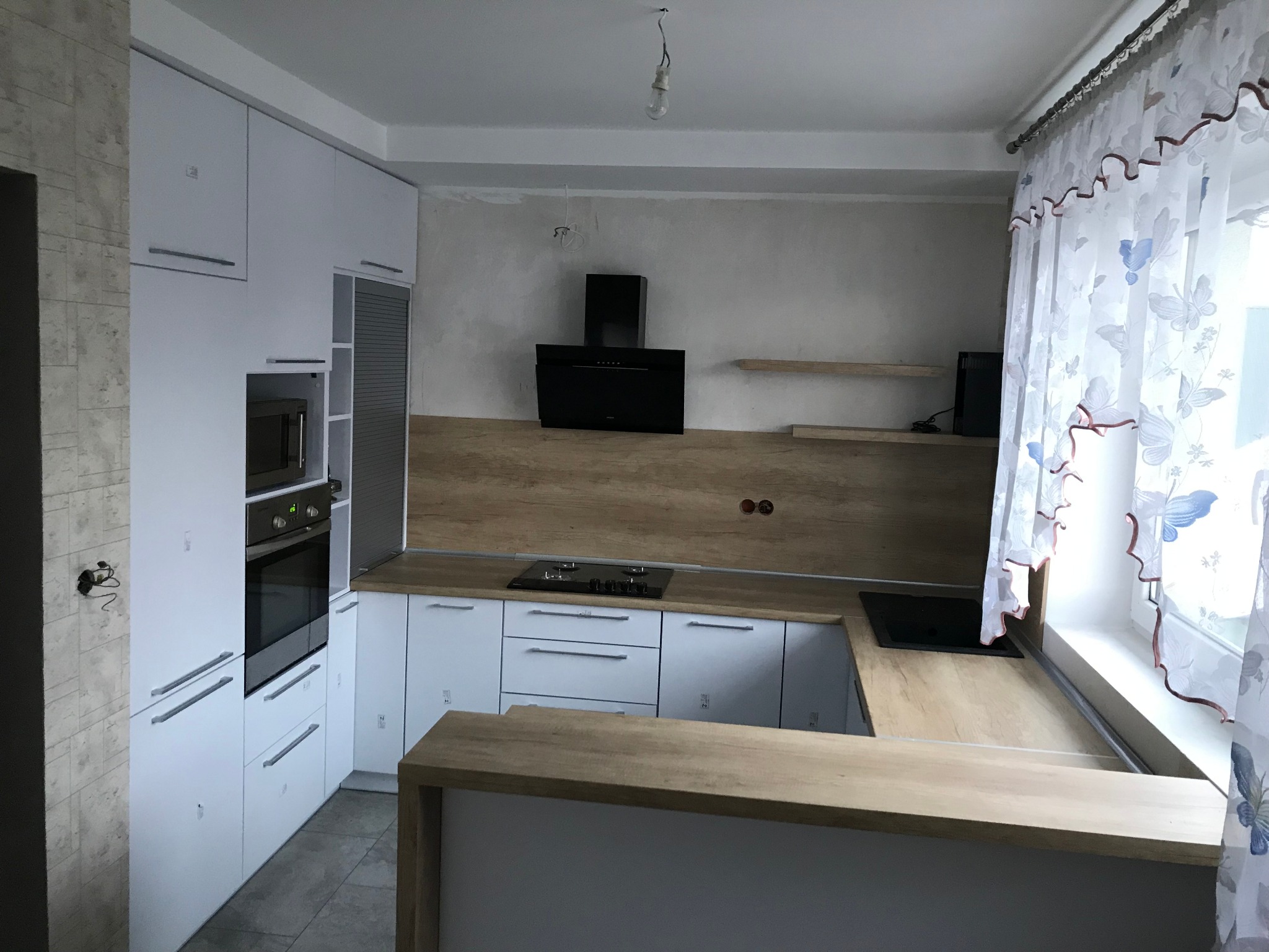 Wnętrze kuchni w trakcie montażu: białe szafki z długimi, srebrnymi uchwytami, drewnopodobny blat, czarny okap, okno z firanką w motyle, surowa ściana z widocznymi przewodami elektrycznymi.