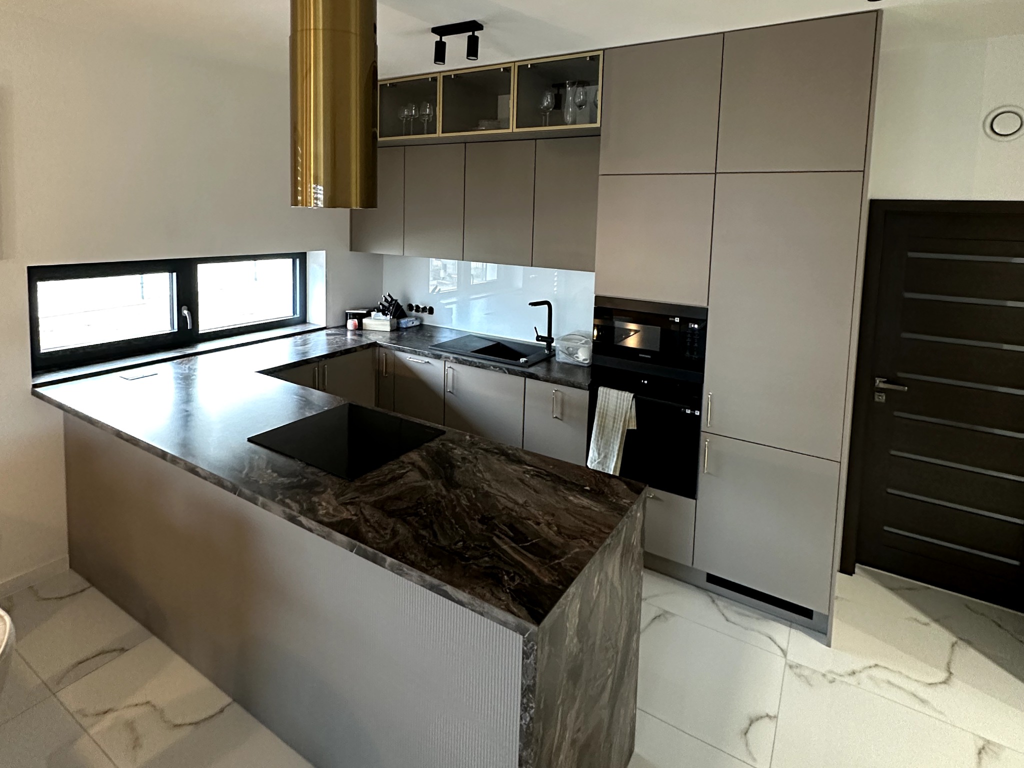 Nowoczesna kuchnia z wyspą i blatem z ciemnego marmuru, złoty okap cylindryczny, szare szafki z podświetlanymi witrynami, czarny zlew i bateria.