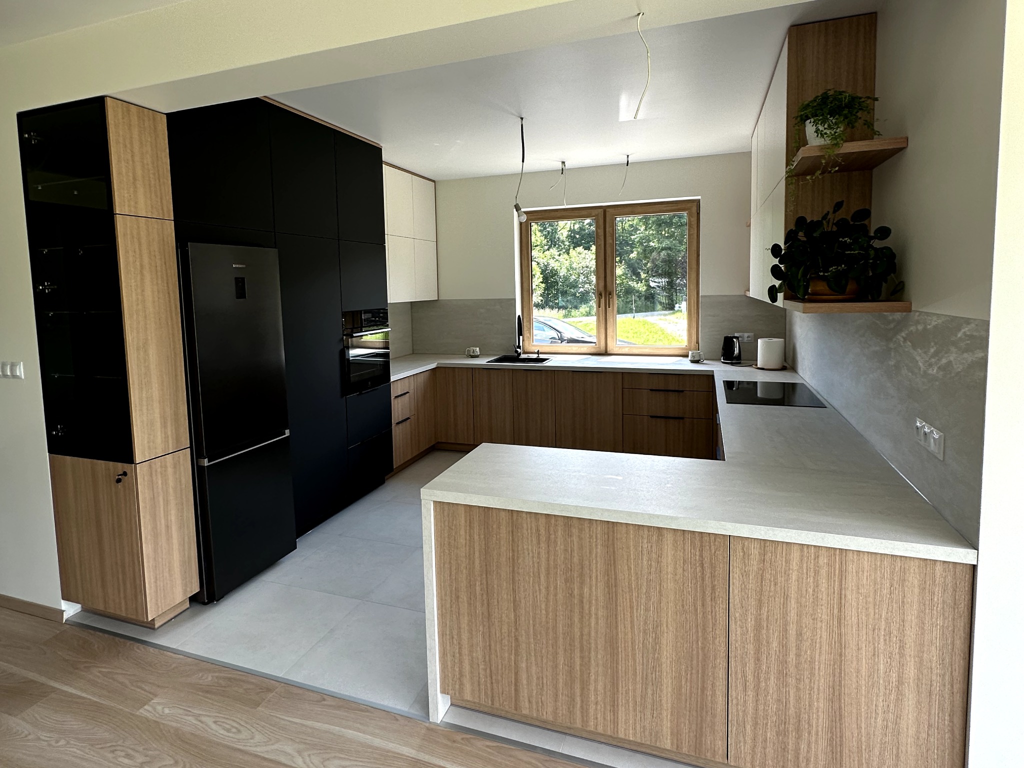 Nowoczesna kuchnia z czarnymi i drewnopodobnymi frontami, wyspą z jasnym blatem i dużym oknem z widokiem na zieleń. Na półkach widoczne rośliny doniczkowe.