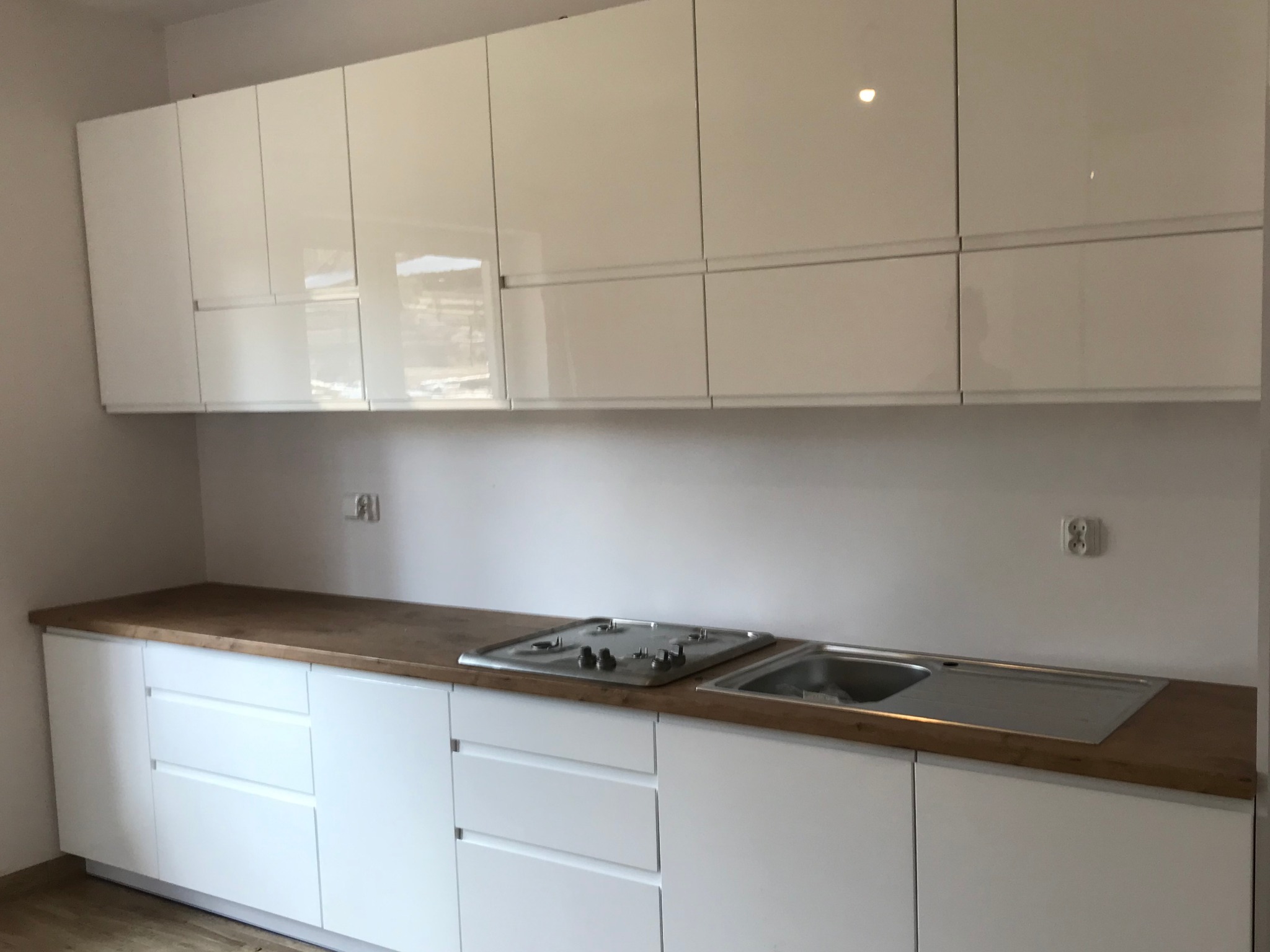 Nowoczesna, biała zabudowa kuchenna z drewnianym blatem, widoczna płyta grzewcza i zlewozmywak, minimalistyczny design, gładkie fronty szafek.