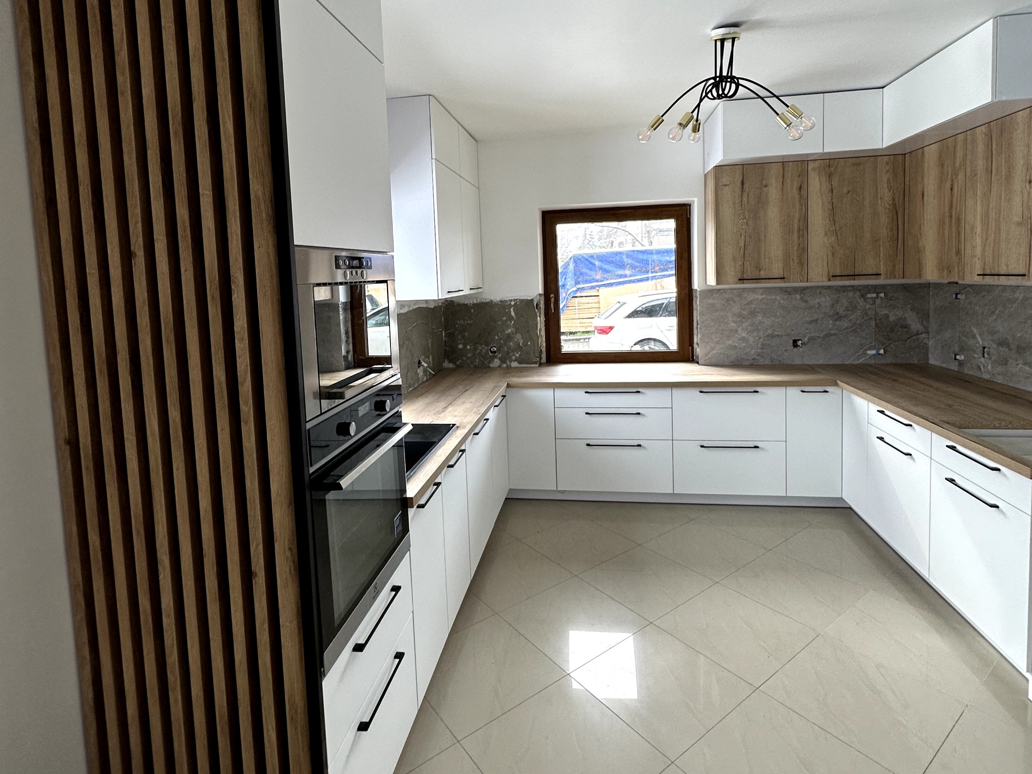 Nowoczesna kuchnia w trakcie montażu: białe szafki z czarnymi uchwytami, drewniany blat, marmurowe wykończenie ściany, drewniane panele dekoracyjne, widoczne okno z zewnątrz i elementy budowlane.