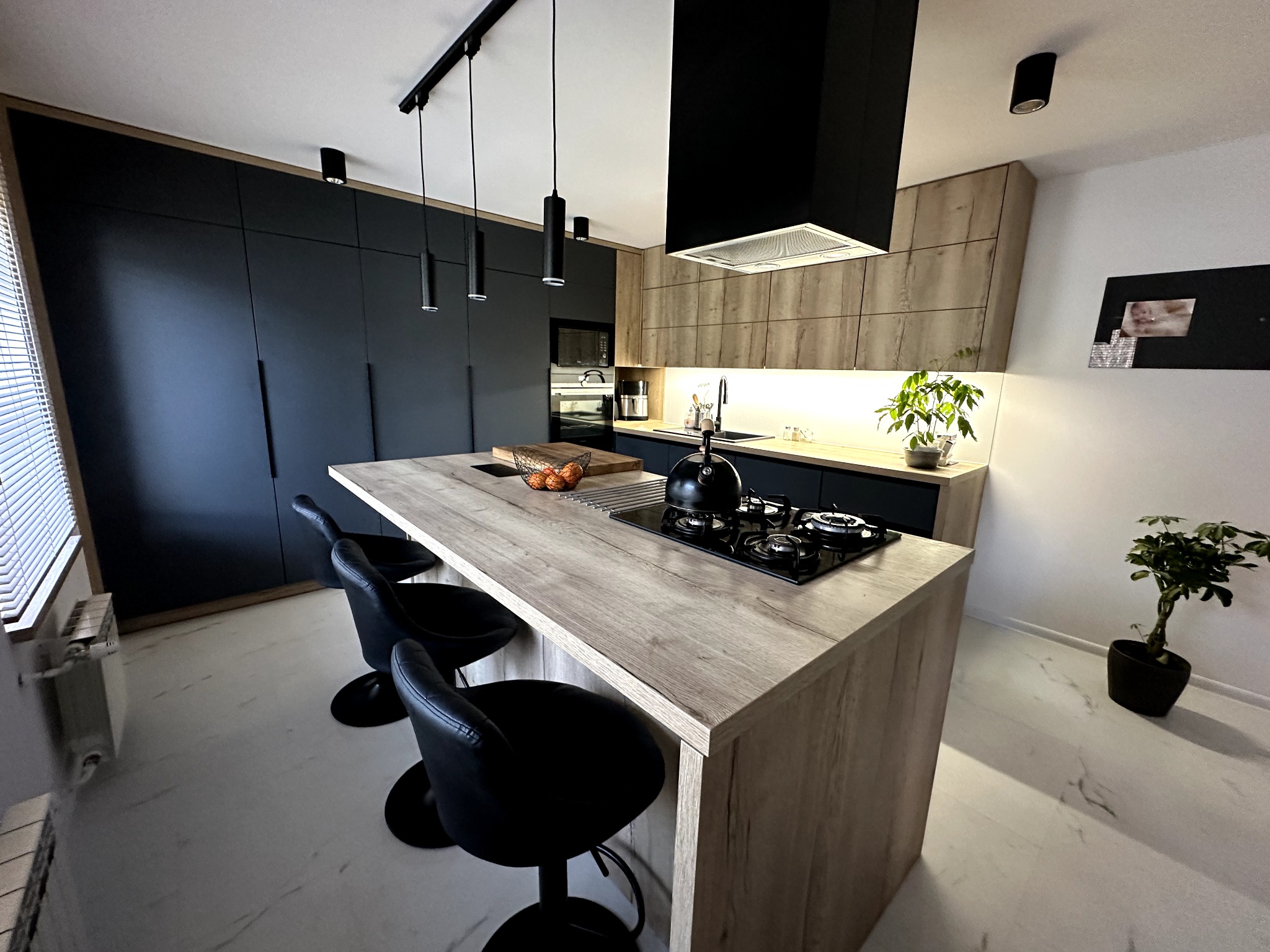 Nowoczesna kuchnia z wyspą i czarnymi hokerami, oświetlenie punktowe i wiszące, fronty szafek w kolorze grafitowym i drewnopodobnym, czarny okap i płyta grzewcza.