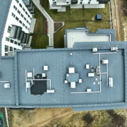Widok z drona na płaski, szary dach budynku wielorodzinnego z licznymi instalacjami technicznymi, w tle widoczne inne budynki i teren z koleinami po pojazdach.
