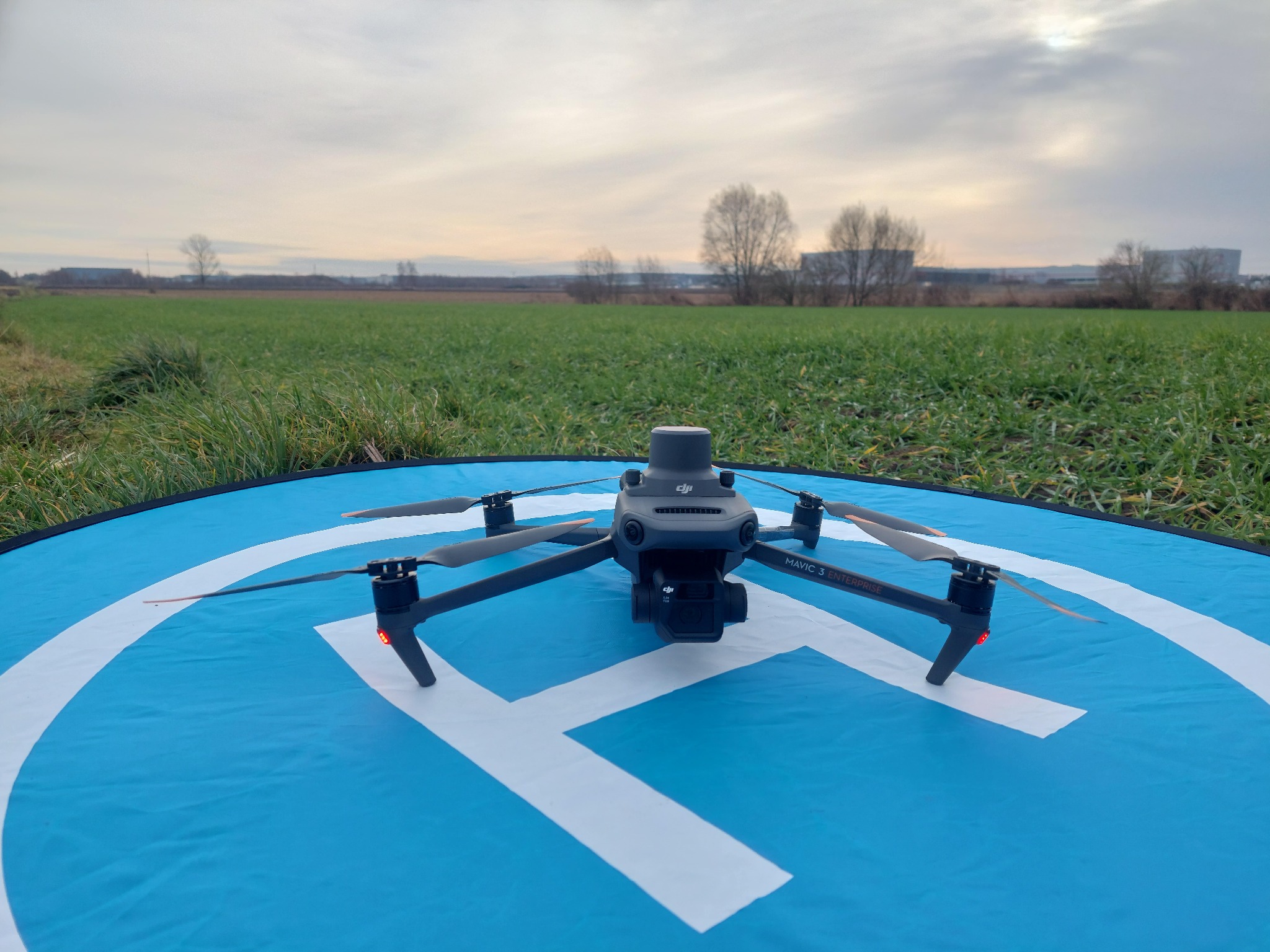 Dron DJI Mavic 3 Enterprise na niebieskiej macie startowej z białym symbolem H, w tle zielone pole i budynki pod pochmurnym niebem.