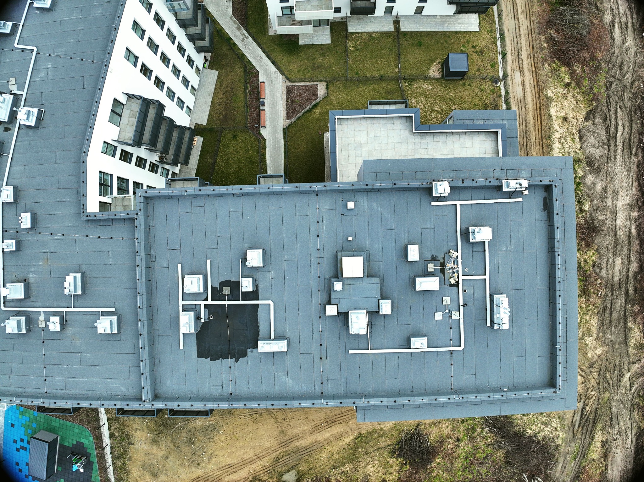 Widok z drona na płaski, szary dach budynku wielorodzinnego z licznymi instalacjami technicznymi, w tle widoczne inne budynki i teren z koleinami po pojazdach.