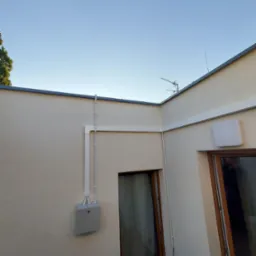 Zewnętrzna instalacja rekuperacji z szarą skrzynką przyłączeniową na elewacji budynku, widoczne białe rury prowadzone wzdłuż ściany i pod dachem, antena telewizyjna na dachu, jasne niebo w tle.