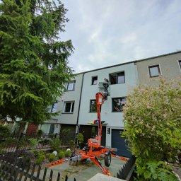 Auto-Klim Konrad Nowak - Pracownik na podnośniku koszowym przed budynkiem mieszkalnym, prawdopodobnie montujący lub serwisujący urządzenie na elewacji.